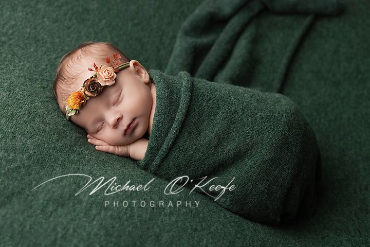 Newborn baby sleeping peacefully wrapped in dark green swaddle blanket during professional photo session.