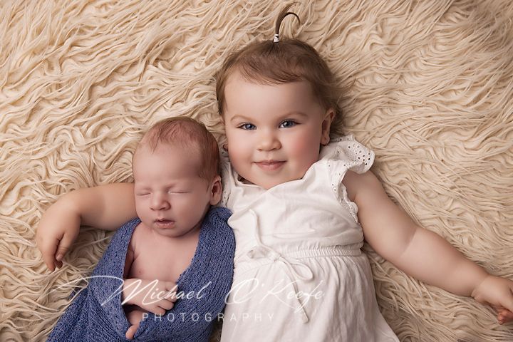 A baby in a blue dress lies on a soft cream-colored blanket with a sweet expression.