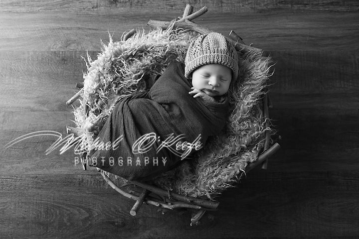 Sleeping newborn in knit hat nestled in cozy nest-like basket with textured bedding, captured in black and white.