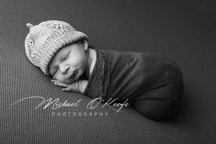 Black and white newborn photo of sleeping baby swaddled in dark wrap wearing knit hat by Michael O'Keefe Photography.