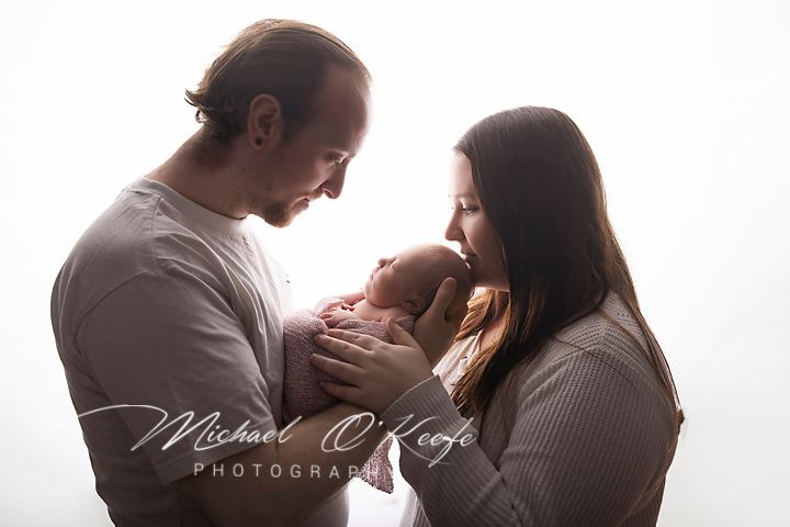 New parents tenderly hold their newborn baby in a soft white studio portrait by Michael O'Keefe Photography.