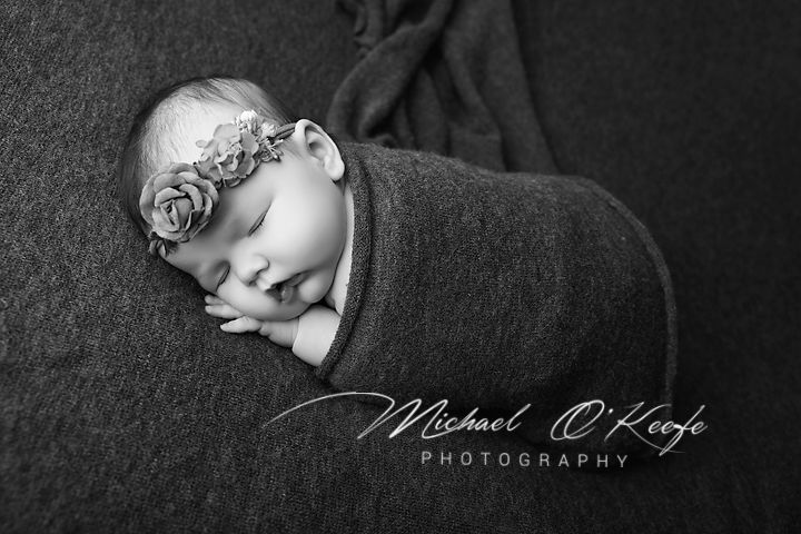 Black and white newborn photography of a sleeping baby wrapped in a blanket with a floral headband by Michael O'Keefe.