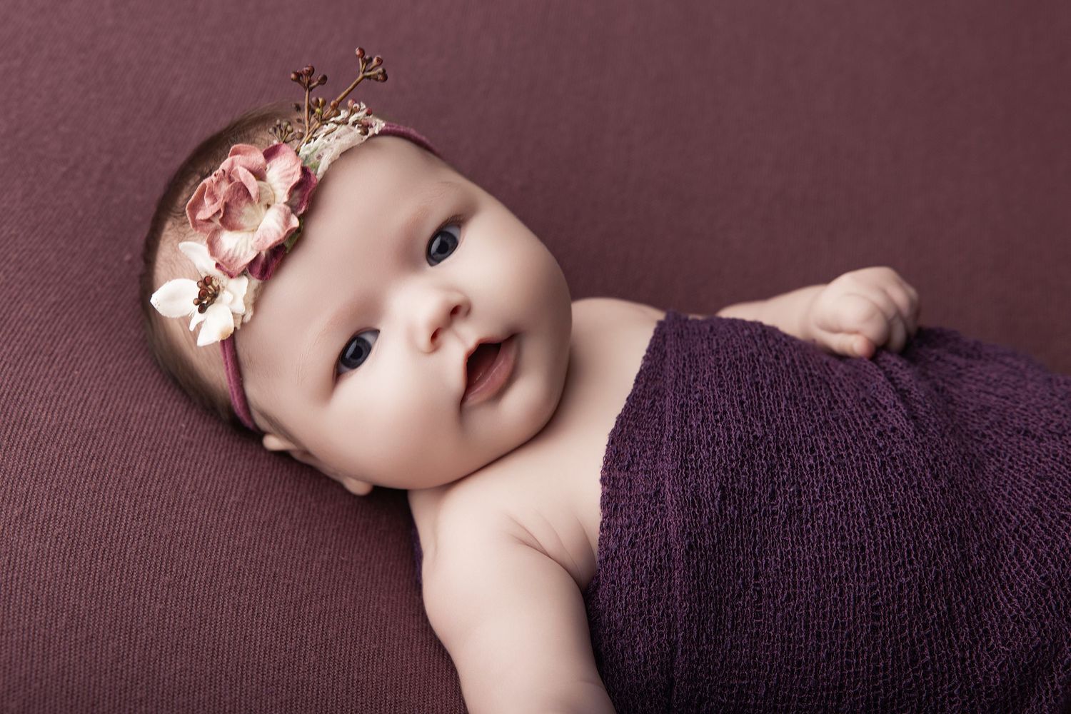 Adorable newborn baby wrapped in purple knit blanket wearing a floral crown headband, lying on a mauve background.