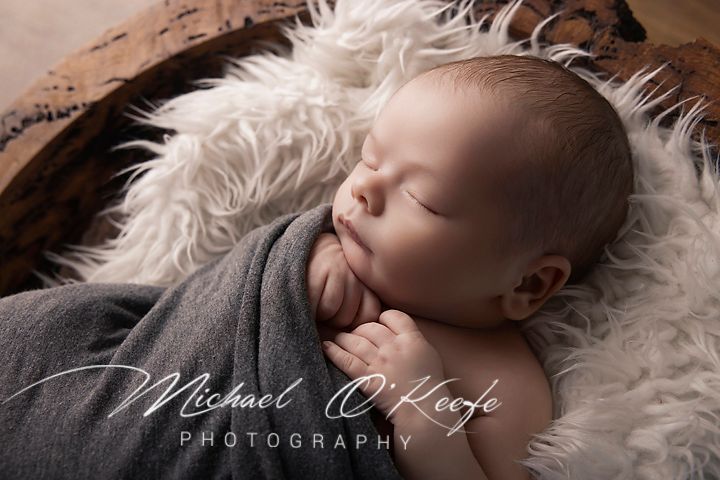 Peaceful newborn baby sleeping wrapped in gray swaddle on fluffy white fur in wooden bowl by Michael O'Keefe Photography.