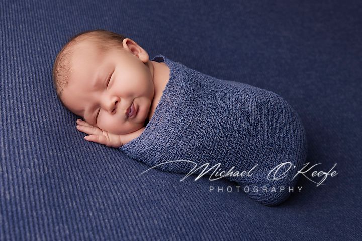 A newborn baby sleeps peacefully on dark blue textured fabric in a professional photography setting.