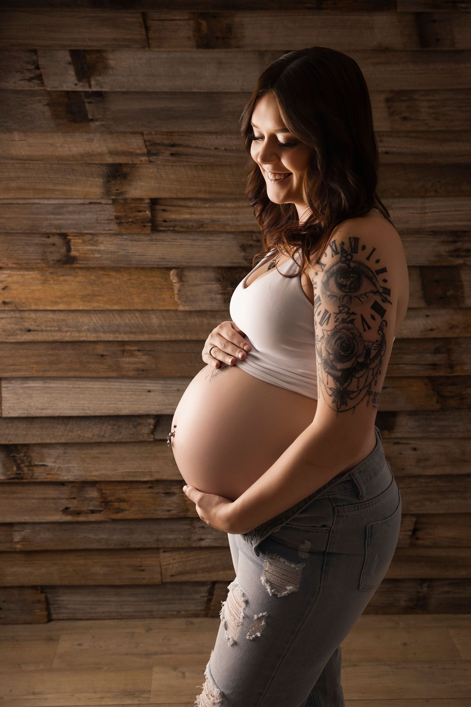 Expectant mother in white tank top and jeans poses against rustic wooden wall, showing floral arm tattoo and baby bump.