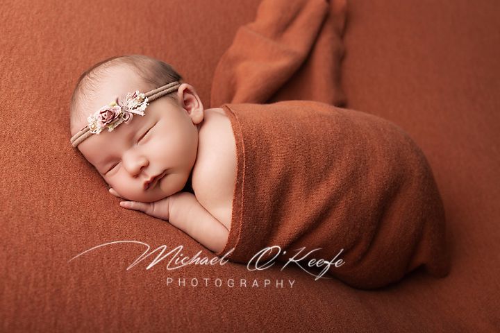 Newborn baby swaddled in rust-orange wrap with floral headband, sleeping peacefully on matching backdrop.