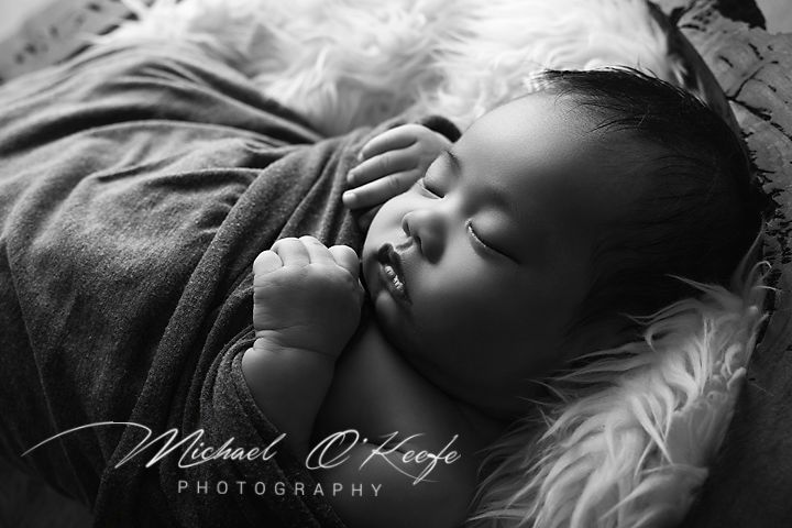 Black and white newborn photography of a sleeping baby wrapped in a soft blanket by Michael O'Keeffe Photography.