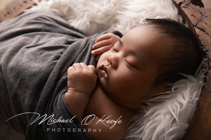Newborn baby sleeping peacefully wrapped in gray swaddle on fur blanket, professional photography by Michael O'Keefe.