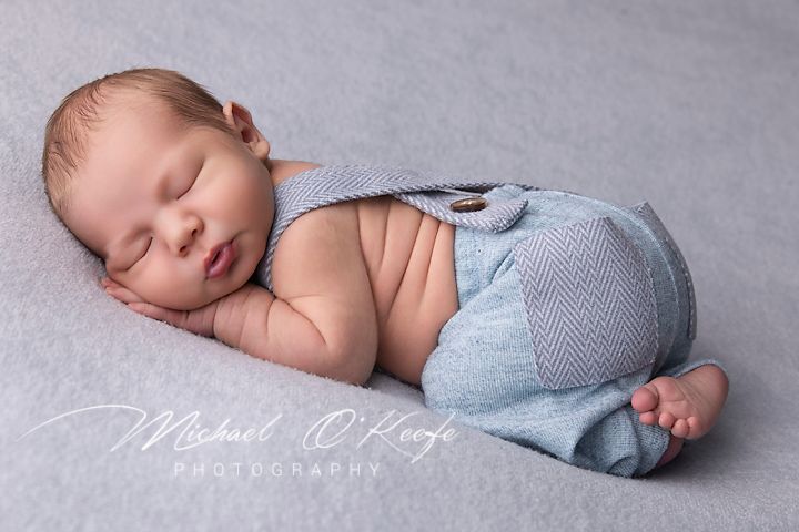 Sleeping newborn wearing light blue shorts lying on side against gray backdrop.