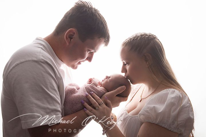 Parents tenderly holding their newborn baby together in a bright, soft-lit professional newborn photography session.