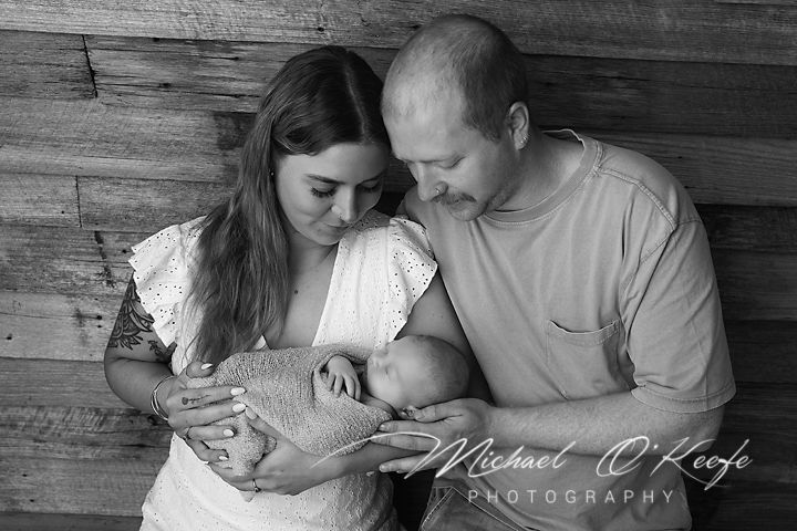A tender black and white portrait of parents with their newborn baby against a rustic wooden wall.