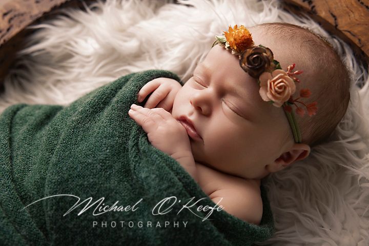 Sleeping newborn wrapped in green blanket on white fur with delicate floral headband in professional portrait.