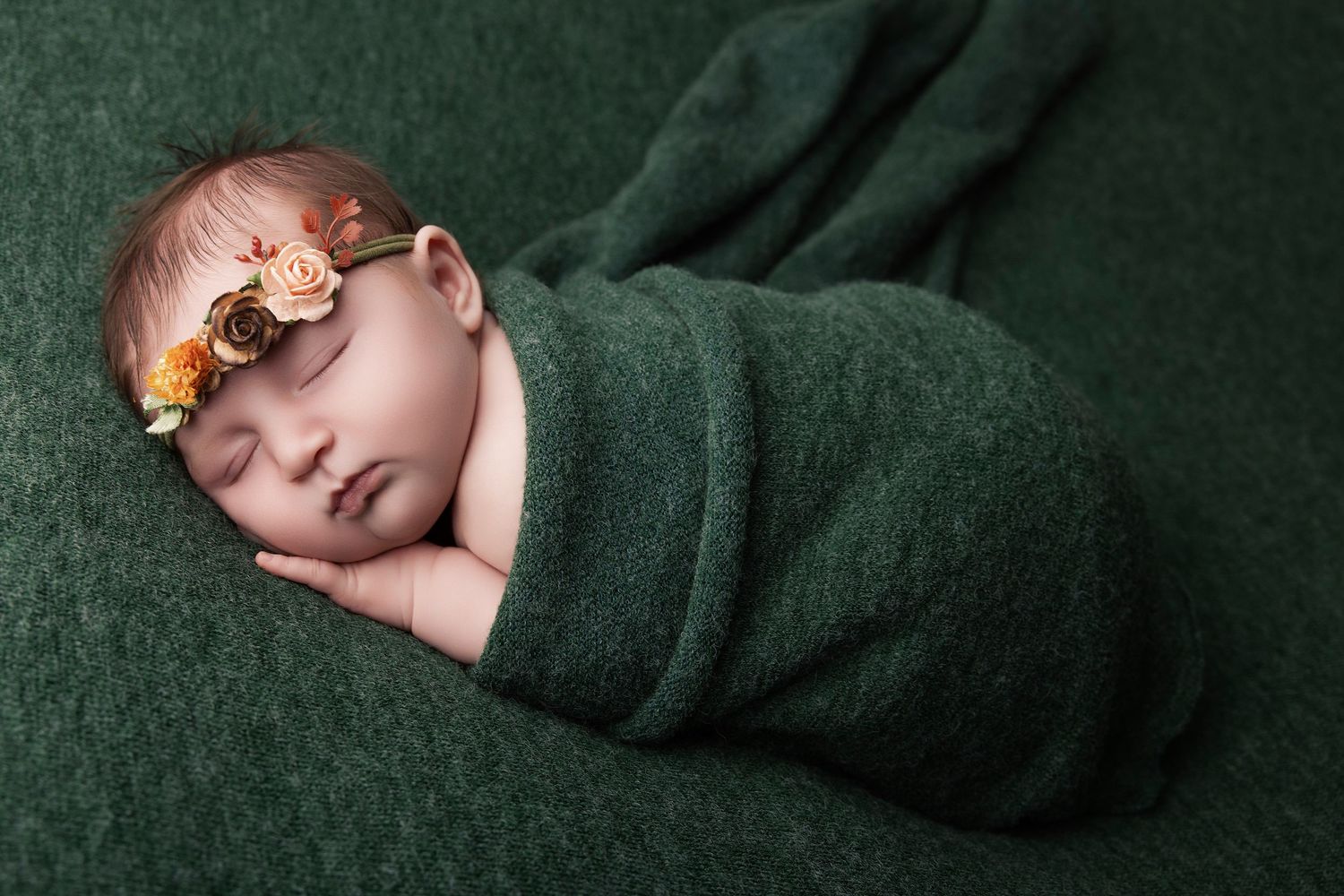 Sleeping newborn baby girl wrapped in a dark green blanket wearing a floral headband, resting on a green textured backdrop.