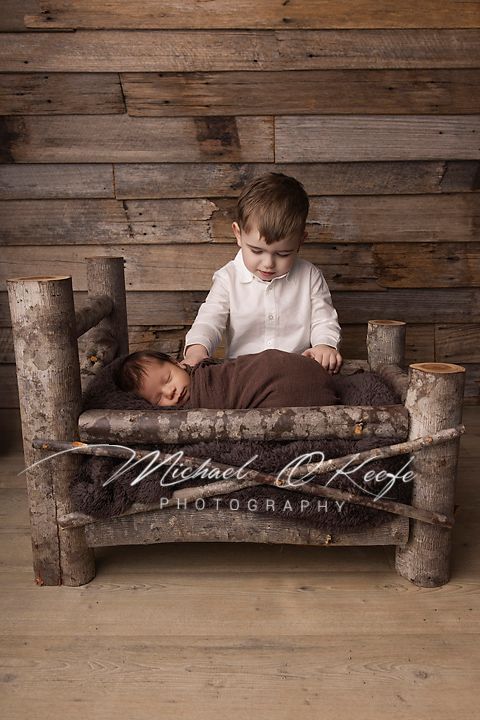 Newborn baby swaddled in brown wrap lying on rustic log bed beside toddler sibling, professional photography.