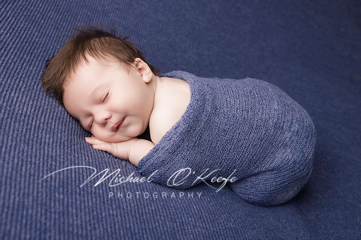 Sleeping newborn wrapped in soft gray swaddle blanket on navy blue textured background.