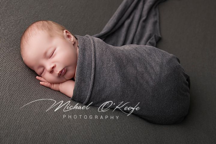 Newborn baby swaddled in gray wrap sleeping peacefully on gray background, photographed by Michael O'Keefe Photography.