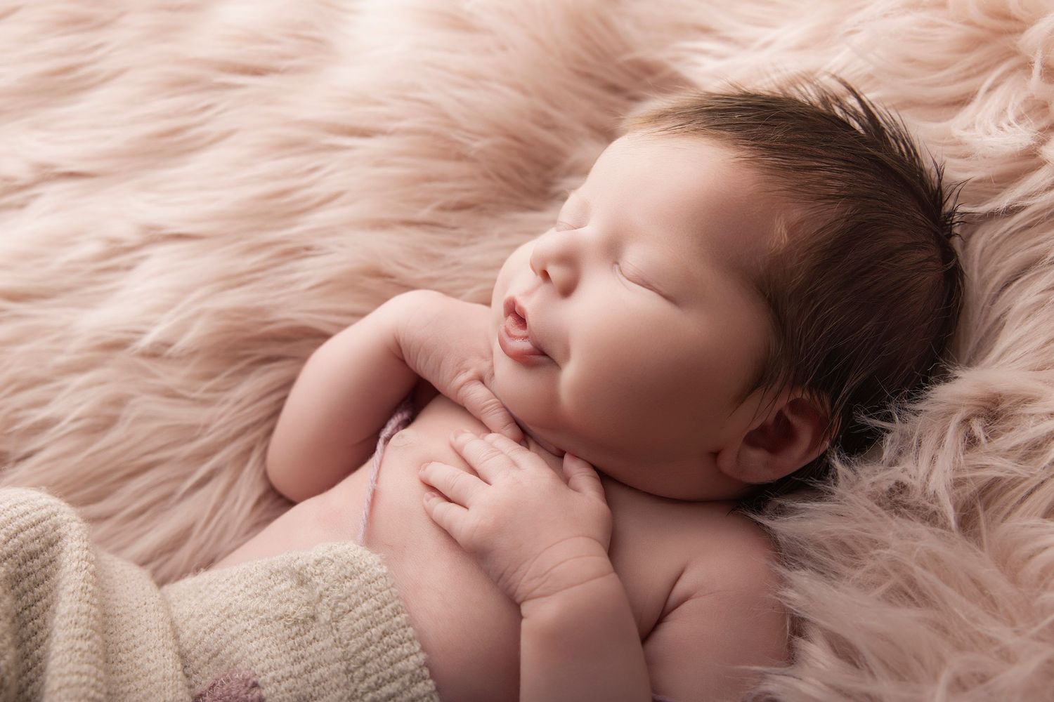 Peaceful newborn baby sleeping on pink fluffy fur blanket with tiny hands folded, soft natural light photography.