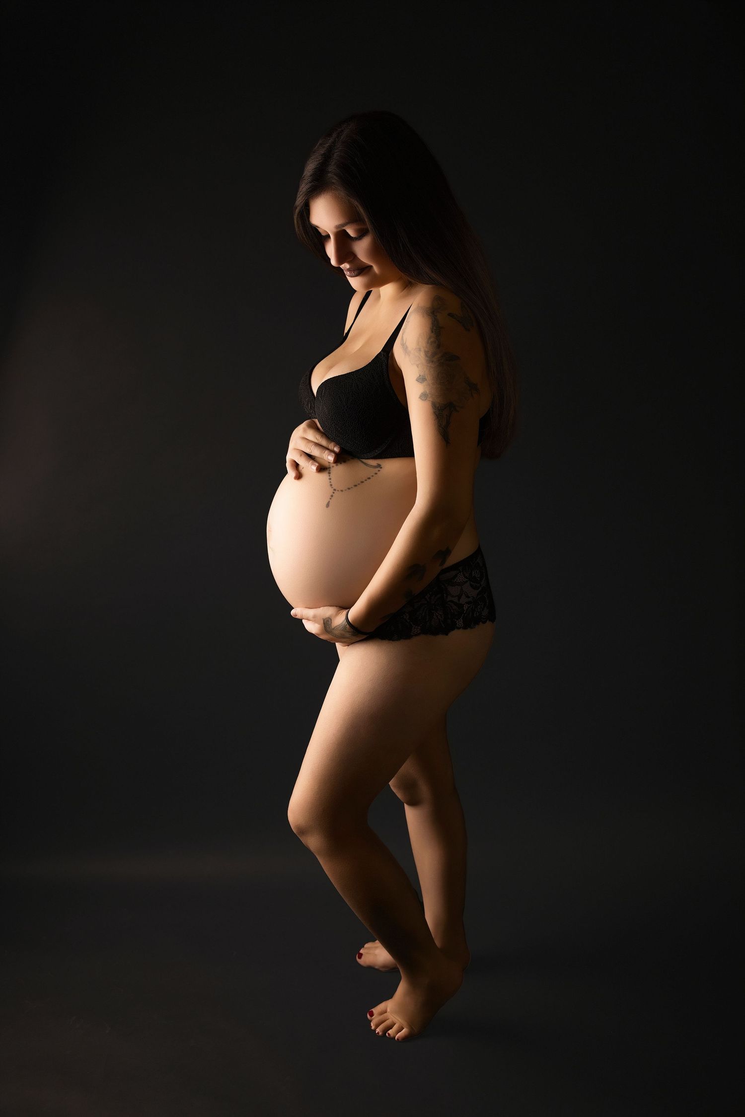 Artistic maternity portrait with dramatic lighting shows an expectant mother in black lingerie against dark background.