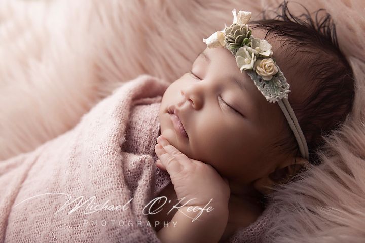 Sleeping newborn baby girl wrapped in pink with floral headband on soft pink fur backdrop.