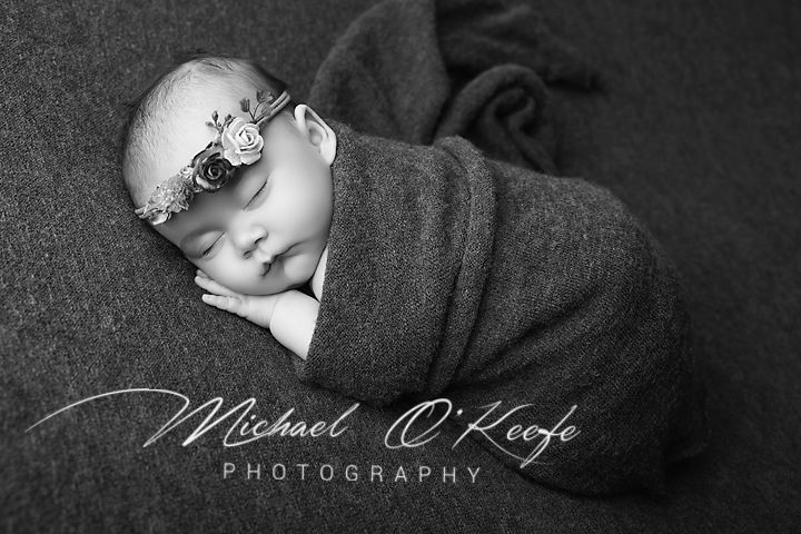 Black and white newborn photo of sleeping baby wrapped in gray knit blanket wearing floral headband by Michael O'Keefe Photography.