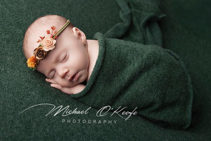 Newborn baby girl sleeping peacefully wrapped in green blanket with floral headband by Michael O'Keefe Photography.