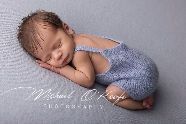 Sleeping newborn baby in blue knit romper on gray backdrop, professional newborn photography by Michael O'Keefe.