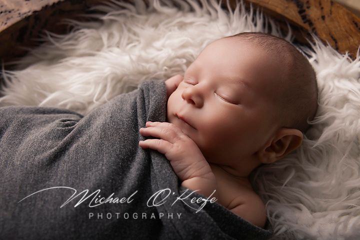 A sleeping newborn baby wrapped in soft gray fabric lies on a plush white fur blanket during a professional photo session.