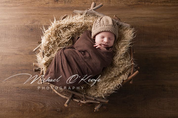Newborn wrapped in brown swaddle sleeping peacefully in rustic nest of hay on wooden backdrop.