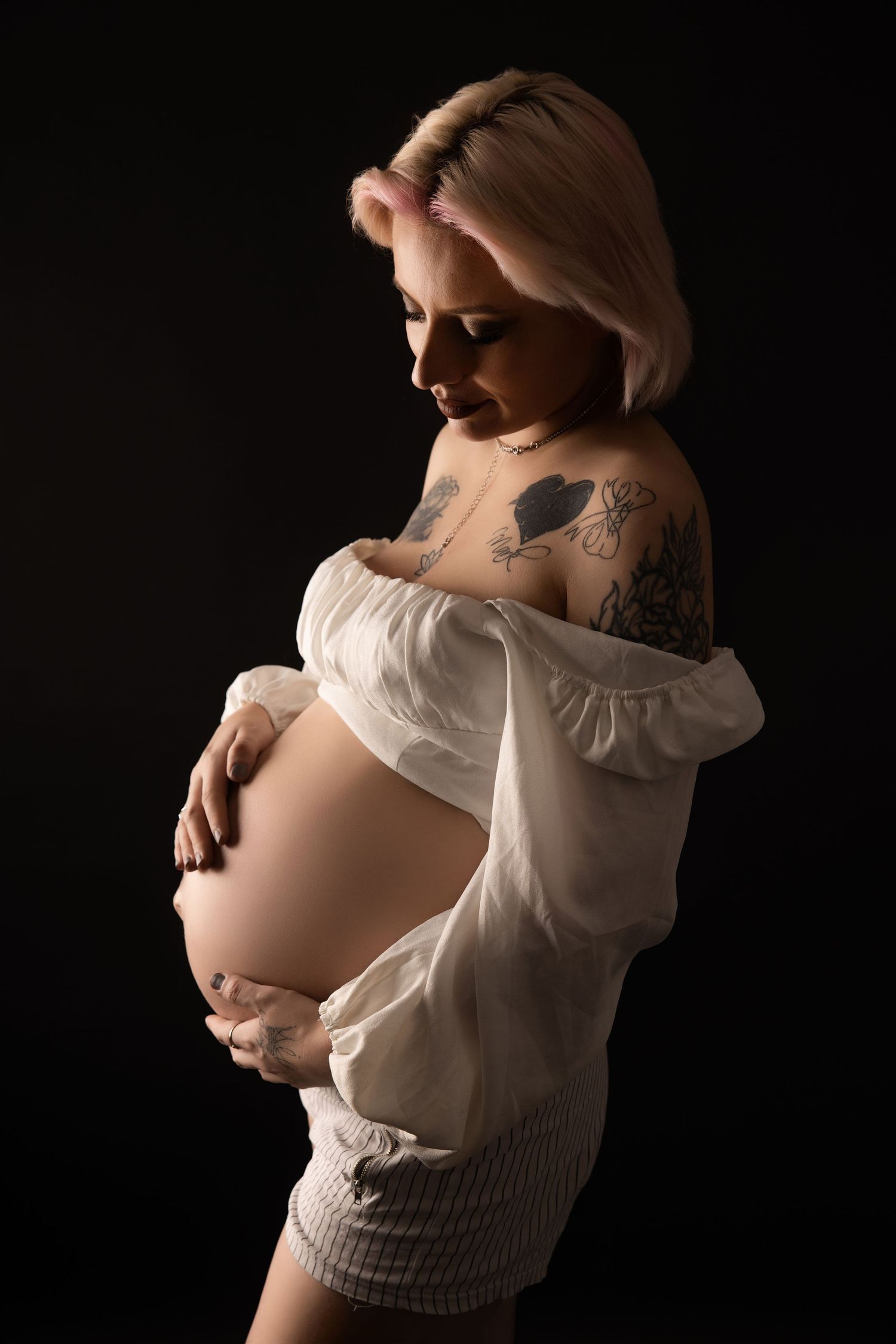 Elegant maternity portraits in white clothing against dark background with dramatic side lighting.