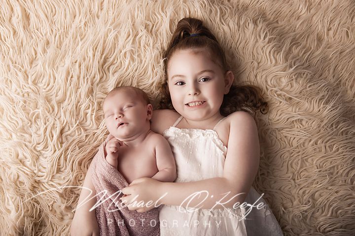 A smiling young girl with a bun hairstyle holds a sleeping newborn baby on a soft cream flokati rug.