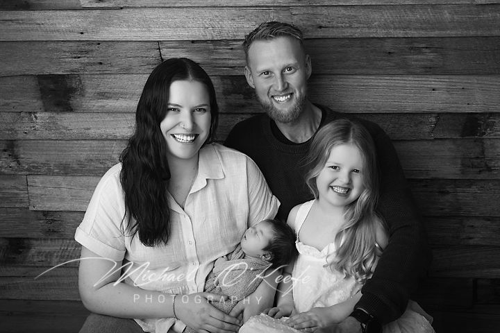 Black and white family portrait with parents holding newborn baby and young daughter, smiling warmly in front of wood backdrop.