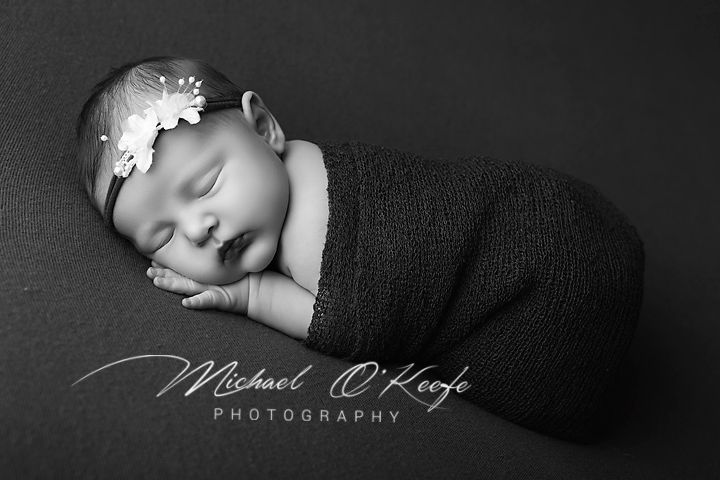 Black and white newborn photography of a sleeping baby wrapped in dark fabric with a floral headband by Michael O'Keefe.