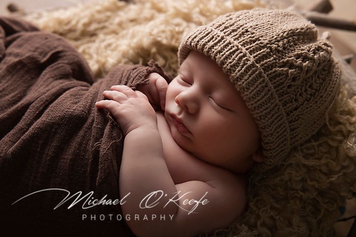 A sleeping newborn baby wearing a brown knit hat lies on soft textured fabric in a professional photography session.
