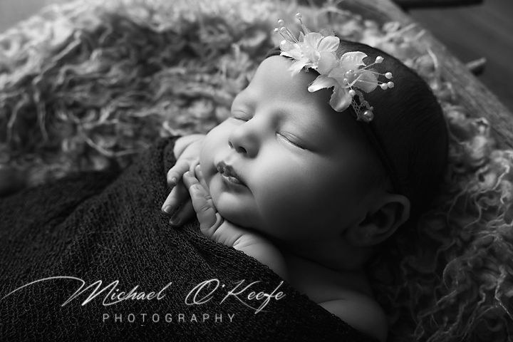 Black and white newborn photography of a sleeping baby girl wearing a delicate floral headband, by Michael O'Keefe.