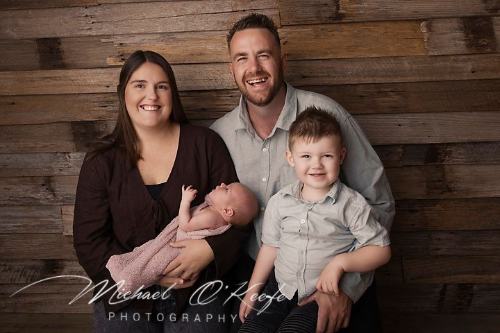 Family portrait with parents holding newborn baby and toddler son, smiling in front of rustic wood wall backdrop.