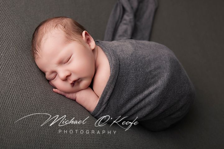 Sleeping newborn baby swaddled in gray wrap posed on gray backdrop by Michael O'Keefe Photography.