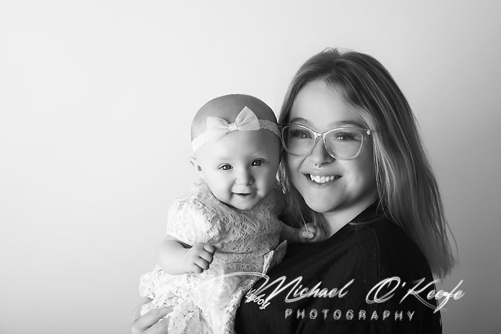 Black and white photo of a young girl with glasses holding a smiling baby wearing a bow headband.