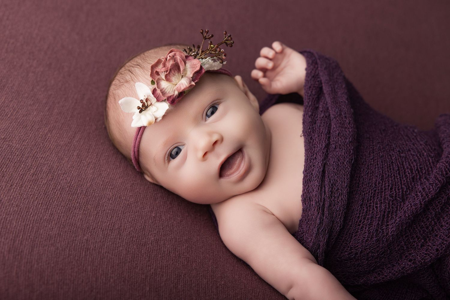 Smiling newborn baby wrapped in purple swaddle wearing a floral crown headband on a mauve background.