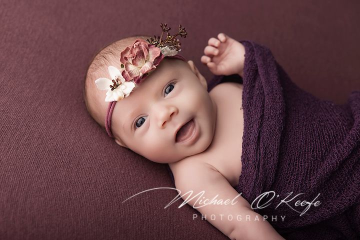 Smiling newborn baby wrapped in purple, wearing a floral crown headband, photographed by Michael O'Keefe Photography.