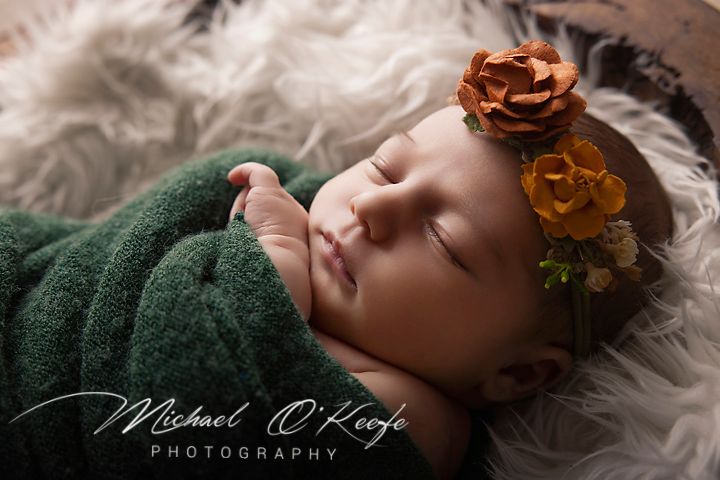 Sleeping newborn baby wrapped in green knit swaddle with floral headband on fluffy white backdrop.