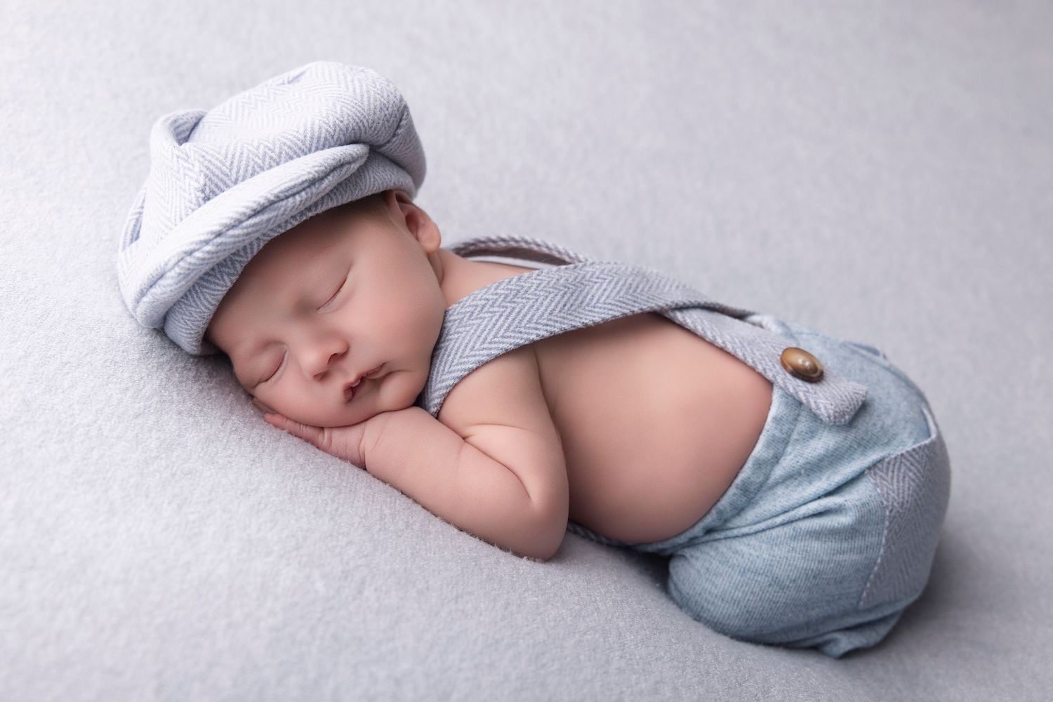Sleeping newborn baby wearing a gray knit cap and suspender shorts outfit on a soft light gray background.
