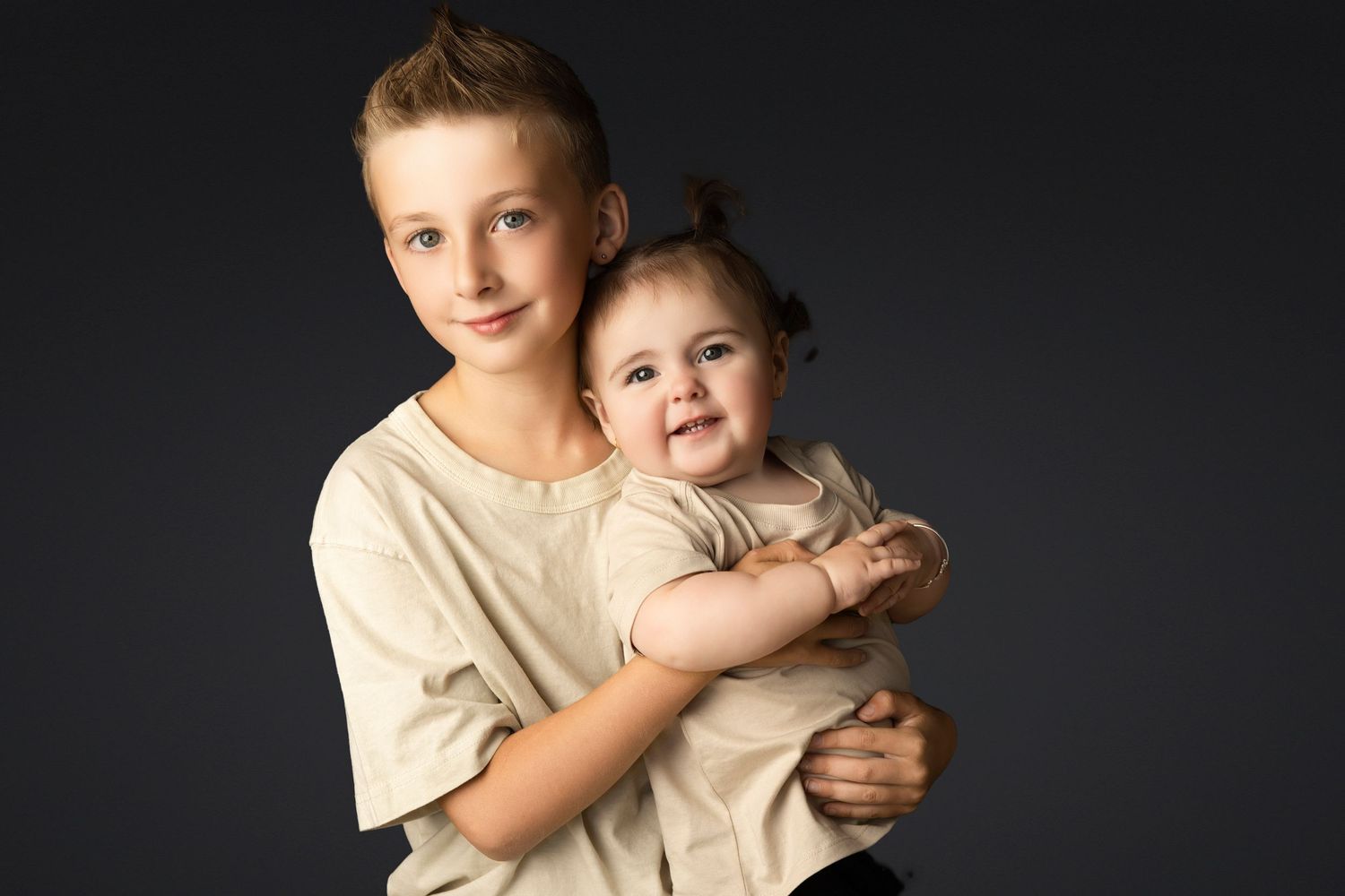 Two siblings in matching beige outfits posing against dark background during portrait session.