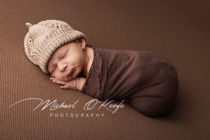 Newborn baby wrapped in brown swaddle wearing knit bear bonnet, sleeping peacefully on brown backdrop.