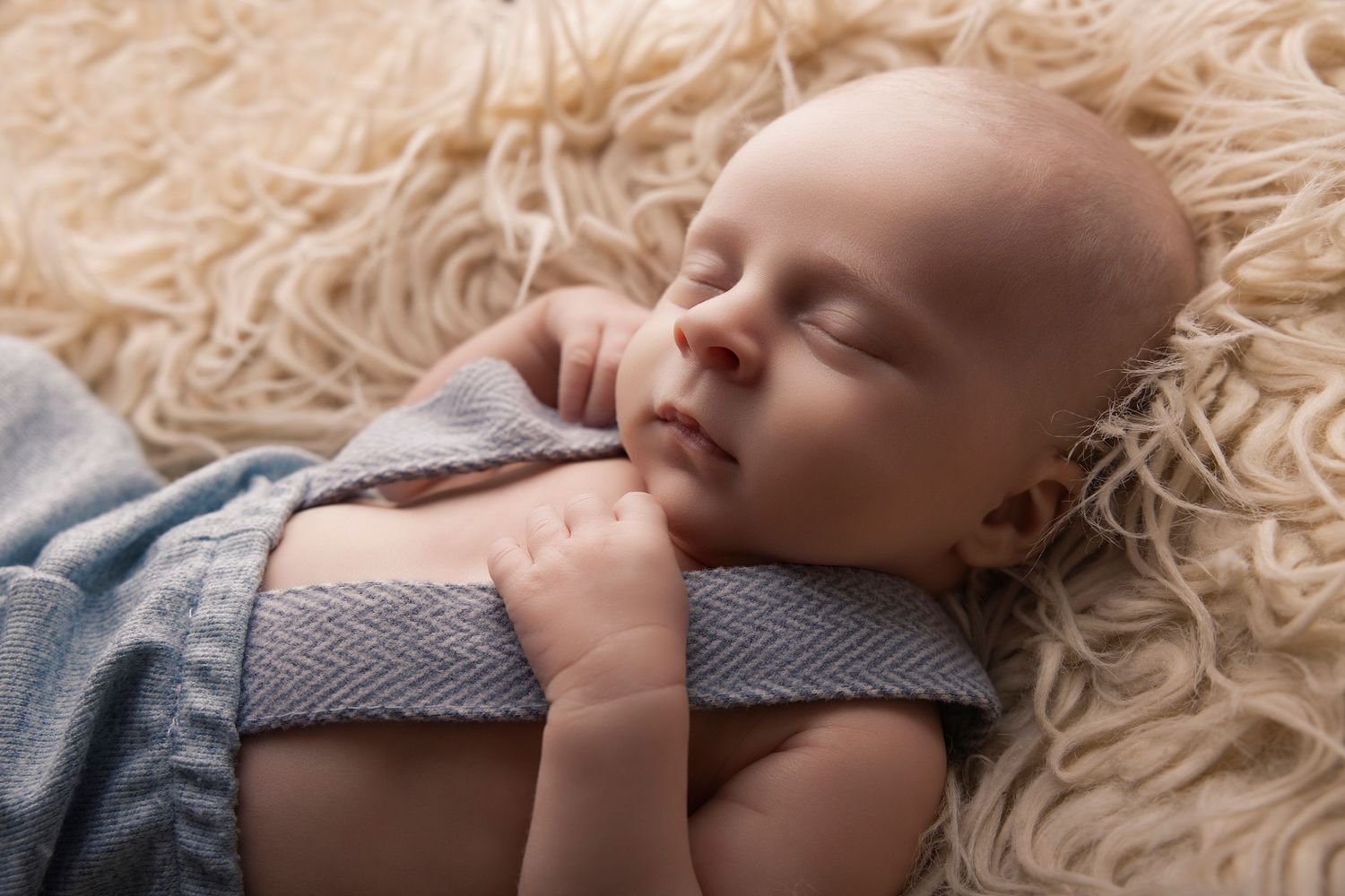 Peaceful newborn baby sleeping on a fluffy cream rug wrapped in a soft blue knit blanket.
