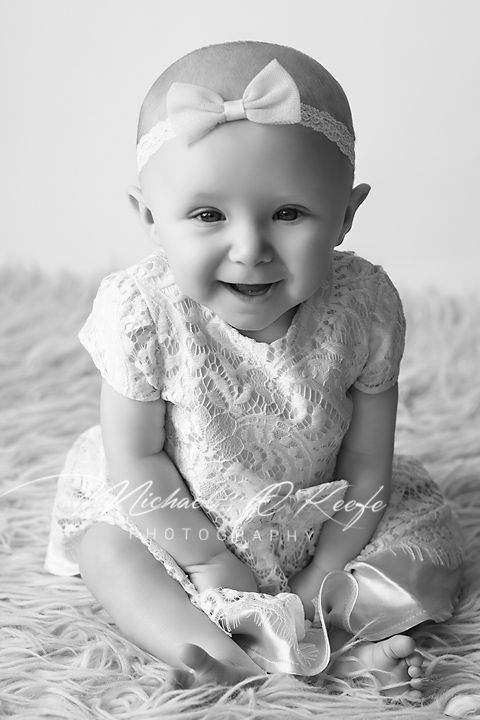 Black and white portrait of a smiling baby girl in lace dress and bow headband sitting on fluffy rug.
