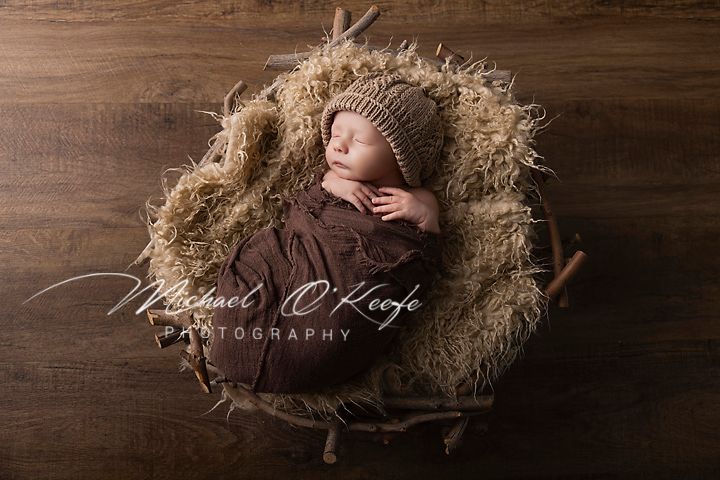 Newborn baby sleeping peacefully in a rustic nest with brown wrap and knit hat, professional photography by Michael O'Keefe.