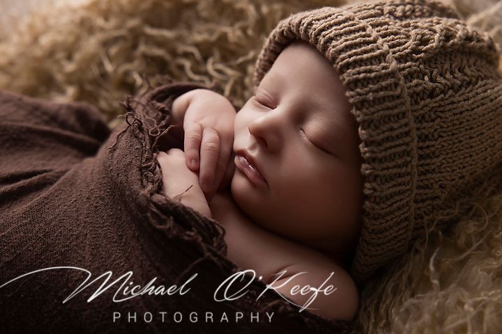 A newborn baby sleeping peacefully on soft brown knit fabric in a professional photography studio setting.