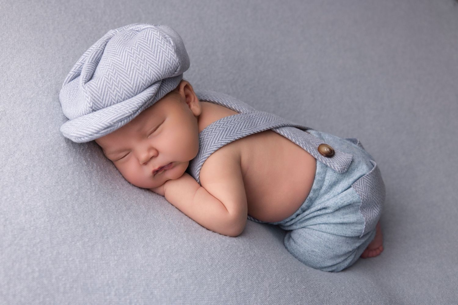 Sleeping baby wearing light gray flat cap and blue suspender shorts lies peacefully on gray backdrop.