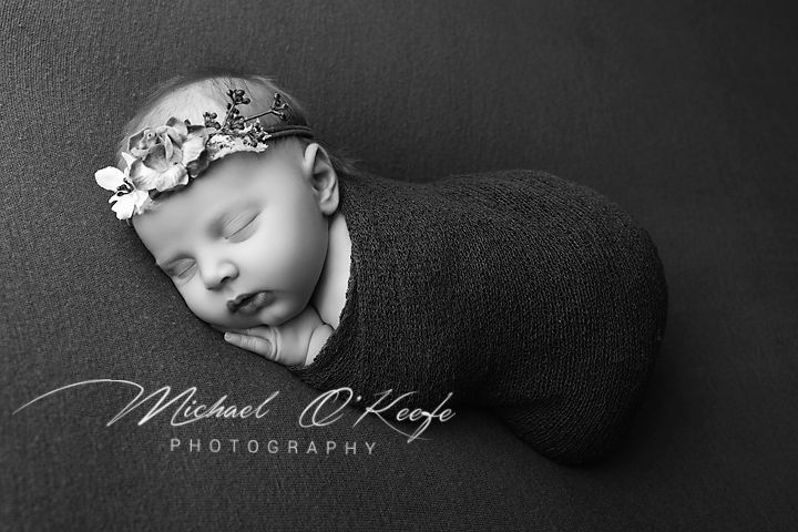 Black and white newborn photo of a sleeping baby swaddled in dark wrap with floral headband by Michael O'Keefe Photography.